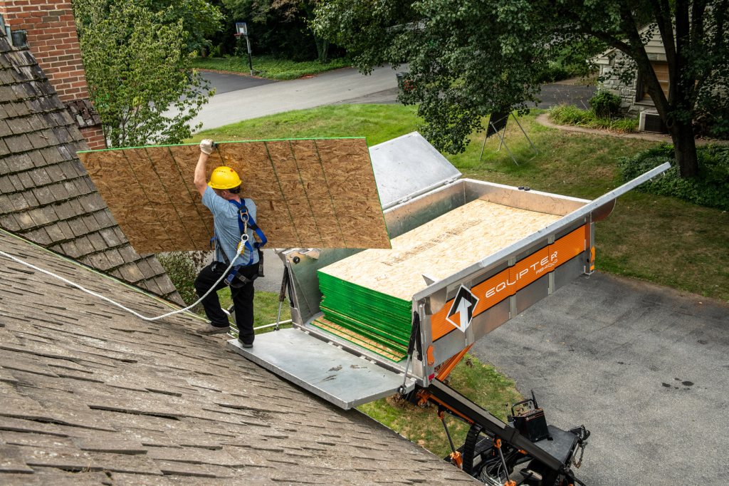 Crew member carrying roof sheathing across a roof toward a debris trailer