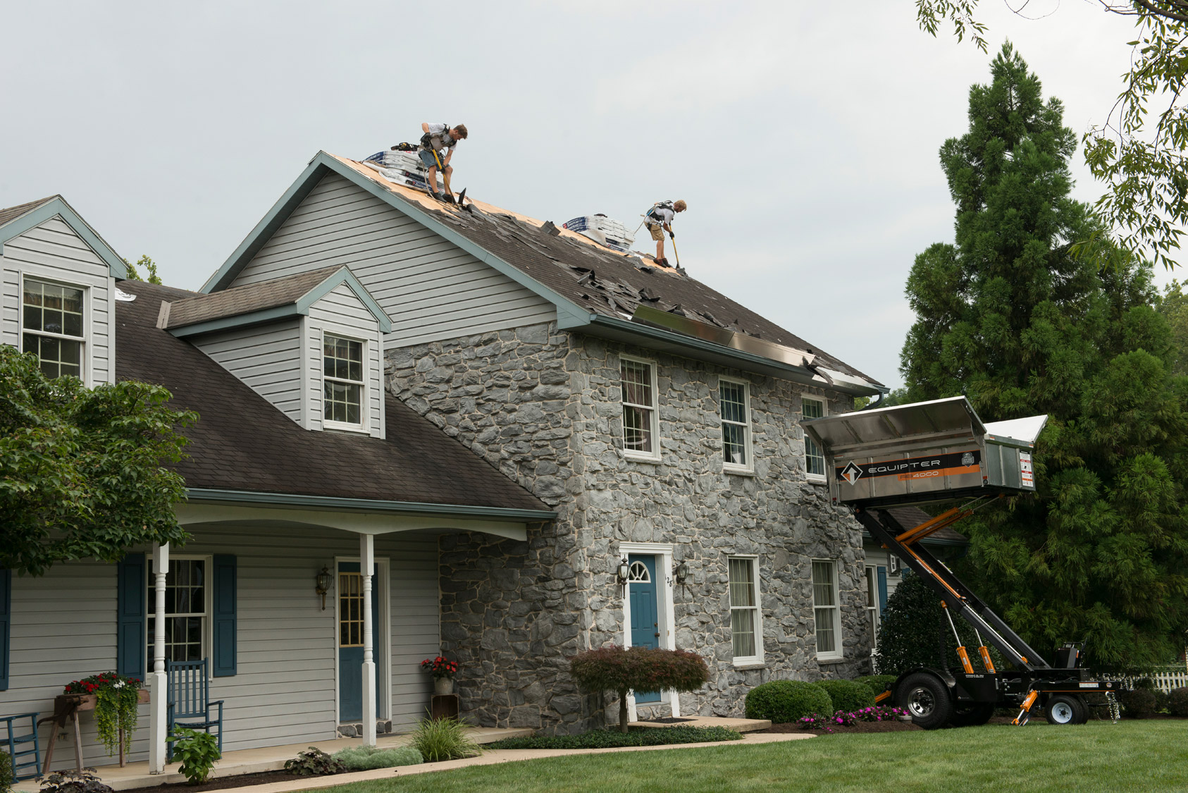 Crew removing old shingles from a stone house, with a debris trailer at the roof edge
