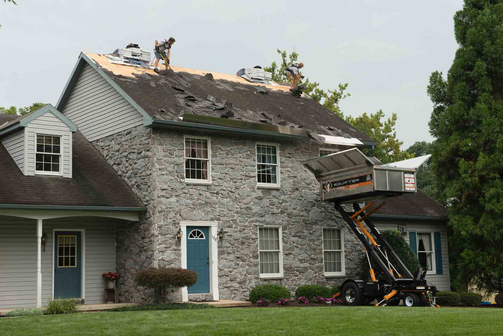 Crew tearing off an old roof on a stone house, with a debris trailer at the eave